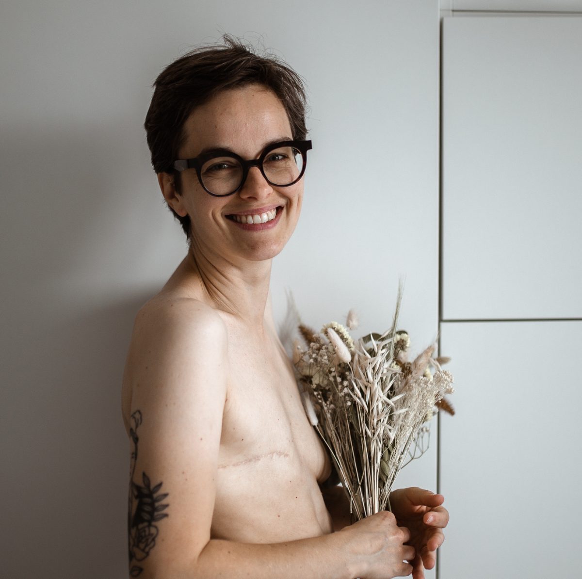 Jeune femme souriante qui cache un de ses seins avec un bouquet et affiche son buste à plat de l'autre côté.